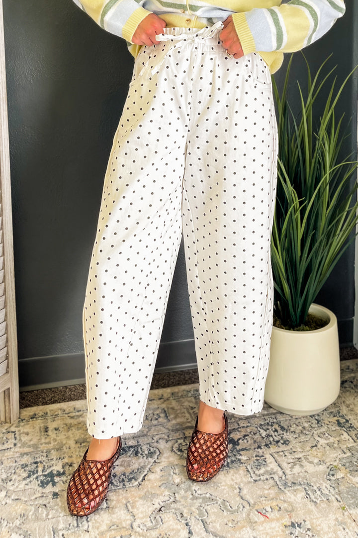 A person wearing white wide-leg cropped pants with a black polka dot pattern, styled with a yellow striped sweater and brown woven sandals, standing indoors next to a potted plant.