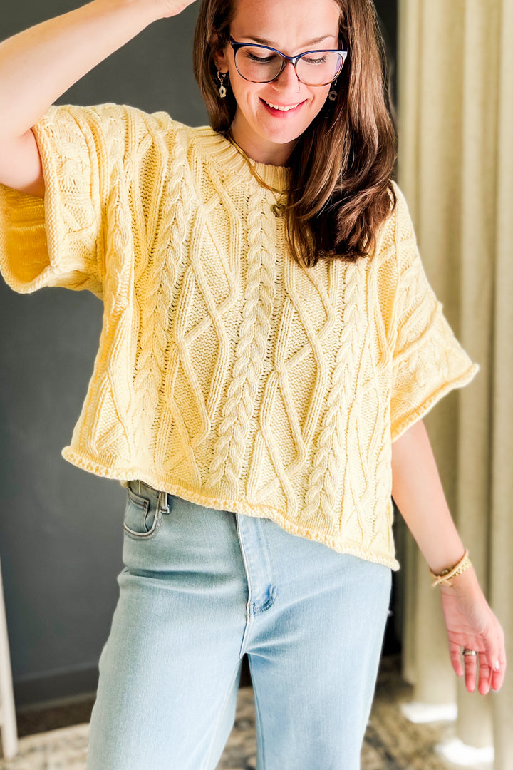 A woman wearing a pale yellow cable-knit short-sleeve top and light blue jeans, standing indoors with a neutral background.
