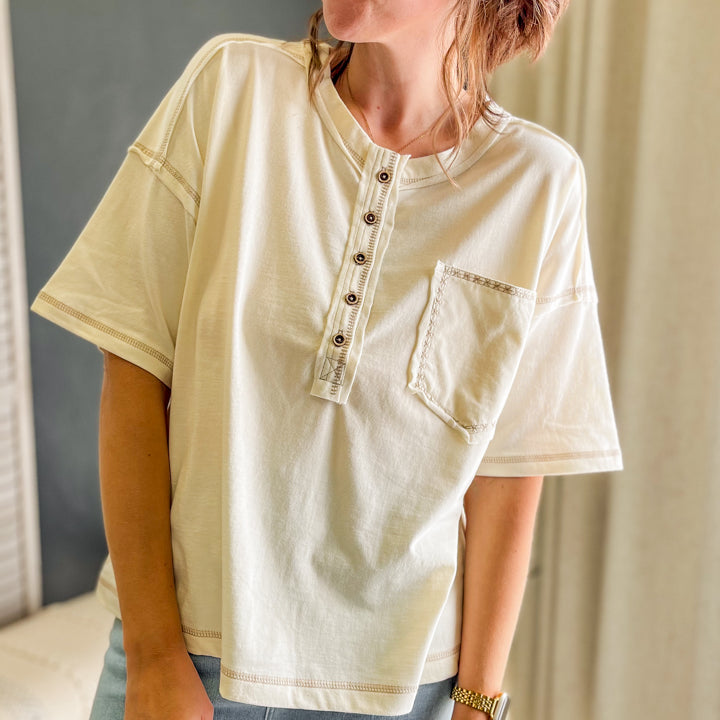 A woman wearing a cream-colored short-sleeve henley top with a button placket and chest pocket, paired with light blue jeans, standing in an indoor setting.