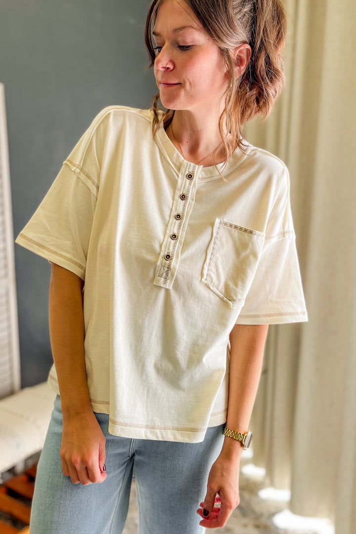 A woman wearing a cream-colored short-sleeve henley top with a button placket and chest pocket, paired with light blue jeans, standing in an indoor setting.