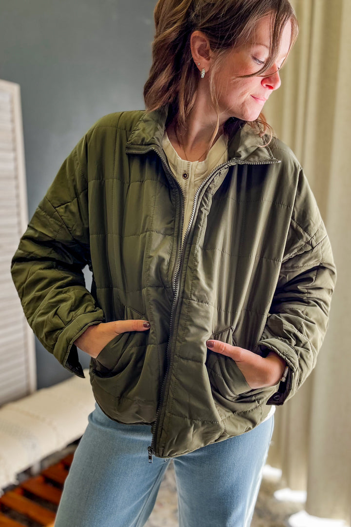 A woman wearing an olive green quilted zip-up jacket over a beige top and light blue jeans, standing indoors with her hands in her pockets.