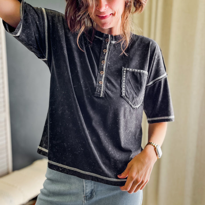A woman wearing a black washed short-sleeve henley shirt with white contrast stitching and light blue jeans, standing indoors near a window with curtains.