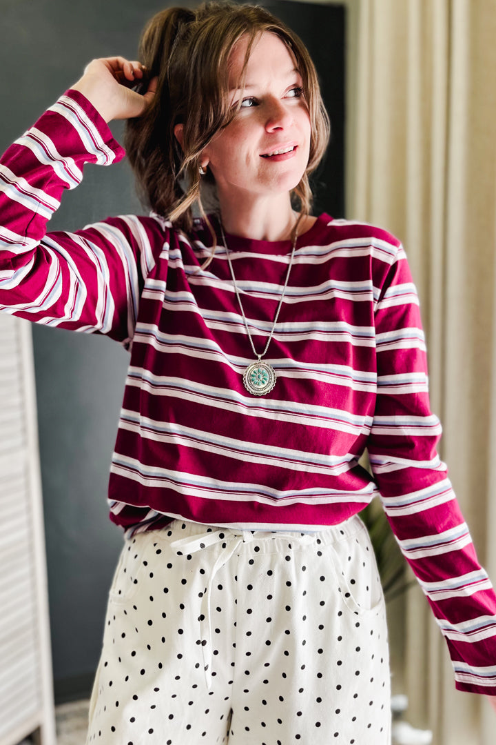 A woman wearing a long-sleeve crewneck shirt with horizontal magenta and white stripes, paired with white pants featuring black polka dots. She is wearing a silver pendant necklace and looking to the side in an indoor setting.