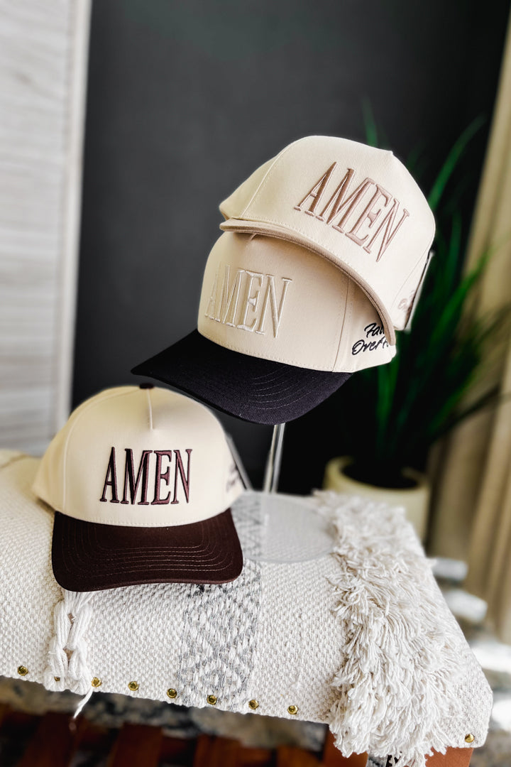 Two beige snapback trucker hats with large embroidered 'AMEN' lettering on the front panels are displayed on a table. One hat has a dark brown brim and the other has a black brim, both styled with a white fringed throw blanket in a cozy indoor setting.