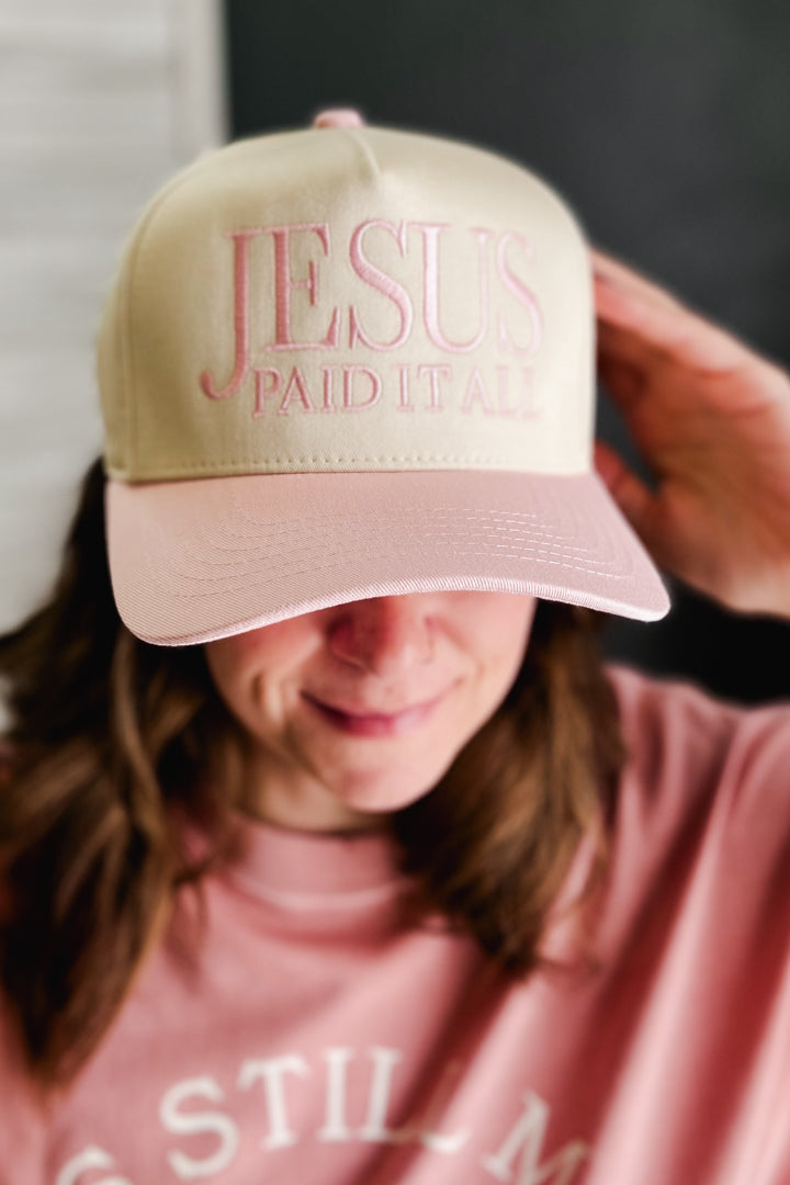 A person wearing a beige baseball cap with pink embroidered text reading 'JESUS PAID IT ALL' and a pink sweatshirt, with the cap covering most of their face.