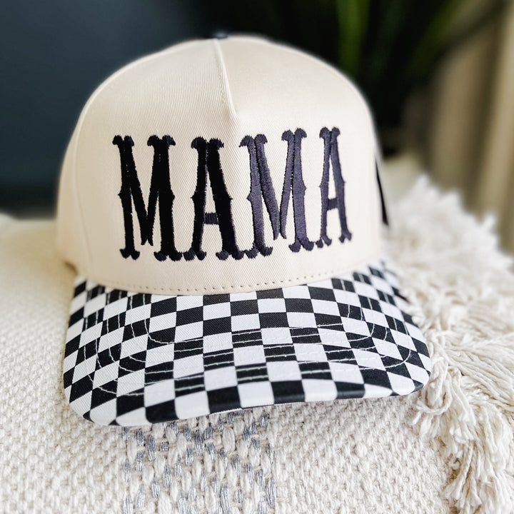 A cream-colored baseball cap with 'MAMA' embroidered in black across the front and a black-and-white checkered pattern on the visor, resting on a textured cream throw blanket.