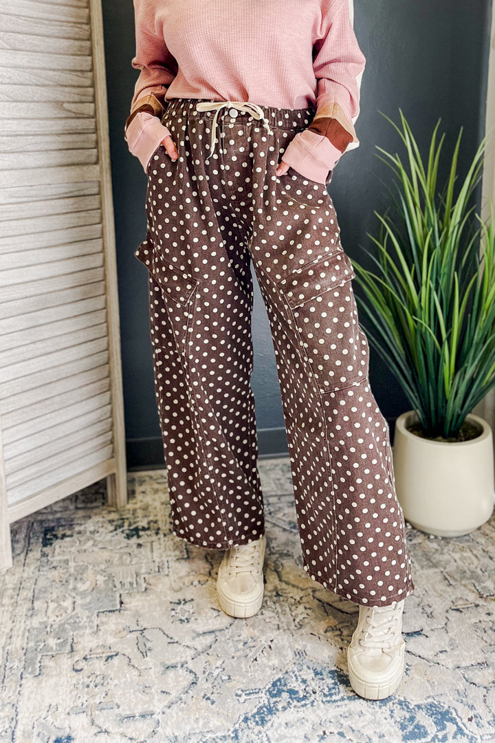 A person wearing brown wide-leg pants with a white polka dot pattern, a pink ribbed sweater, and cream sneakers, standing in an indoor setting with a potted plant and a slatted screen.