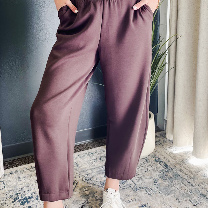 A person wearing loose, wide-leg cropped pants in a dusty mauve or plum-brown color, paired with a white top and white sneakers, standing indoors on a patterned rug.