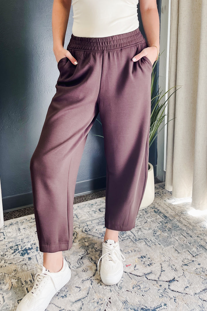A person wearing loose, wide-leg cropped pants in a dusty mauve or plum-brown color, paired with a white top and white sneakers, standing indoors on a patterned rug.