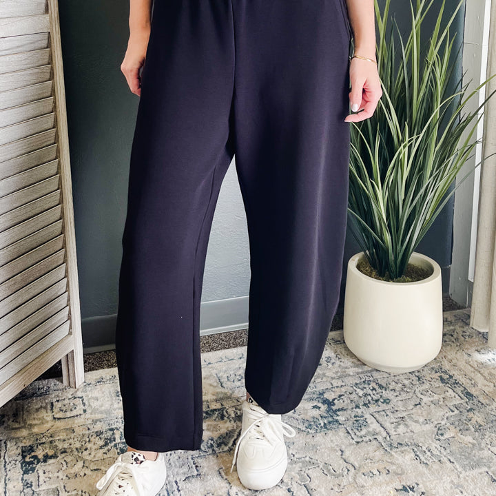 A person wearing dark navy wide-leg sweatpants with an elastic waistband, paired with white sneakers, standing in an indoor setting with a patterned rug and a potted plant.