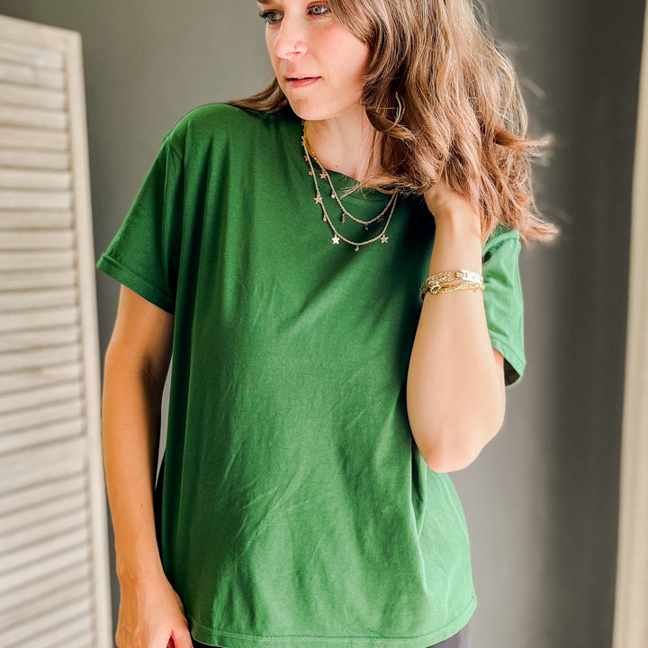 A woman wearing a green short-sleeve t-shirt and dark gray pants, standing indoors against a dark gray wall.