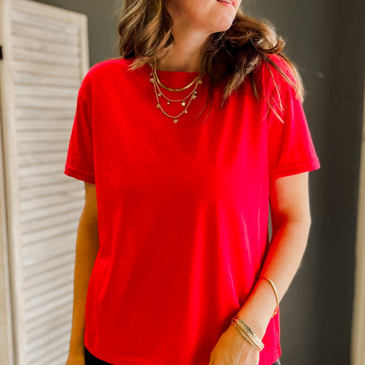 A woman wearing a bright red short-sleeve crewneck t-shirt and dark pants, standing in an indoor setting with a dark gray wall and a white louvered door in the background.