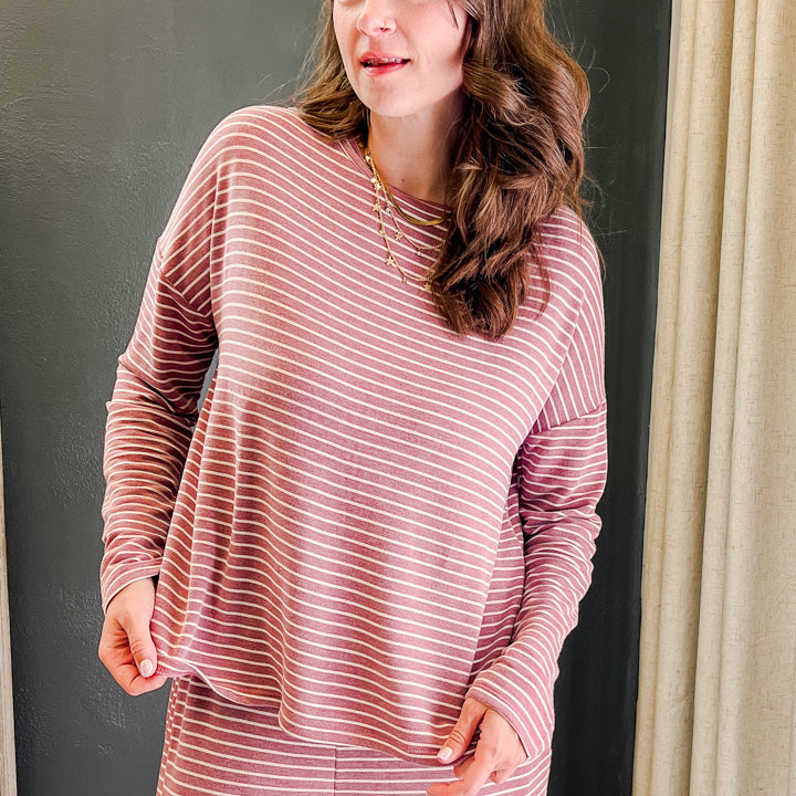 A woman wearing a matching two-piece pajama set with a dusty rose and white striped pattern, standing in front of a dark gray wall and a beige curtain.