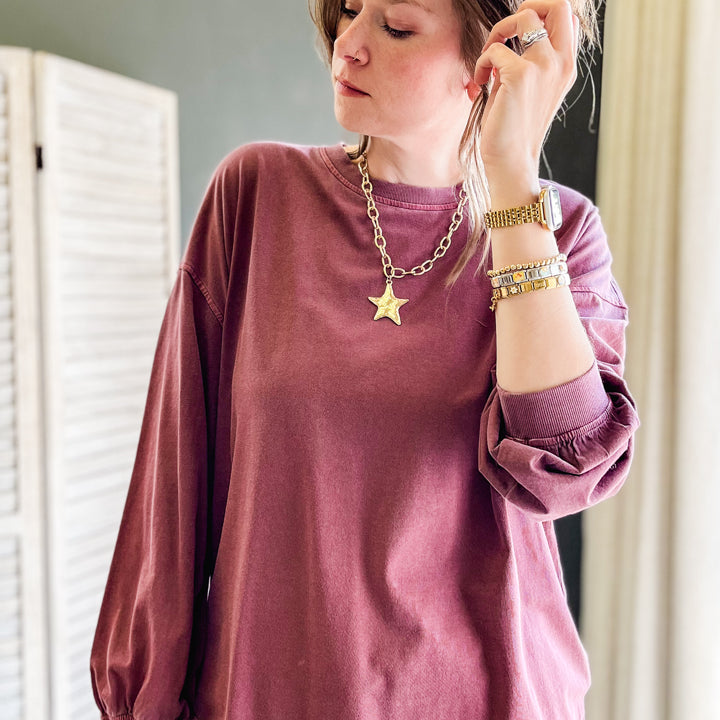A woman wearing a washed mauve long-sleeve t-shirt and blue jeans, accessorized with a gold star pendant necklace and bracelets, standing indoors next to a white folding screen.