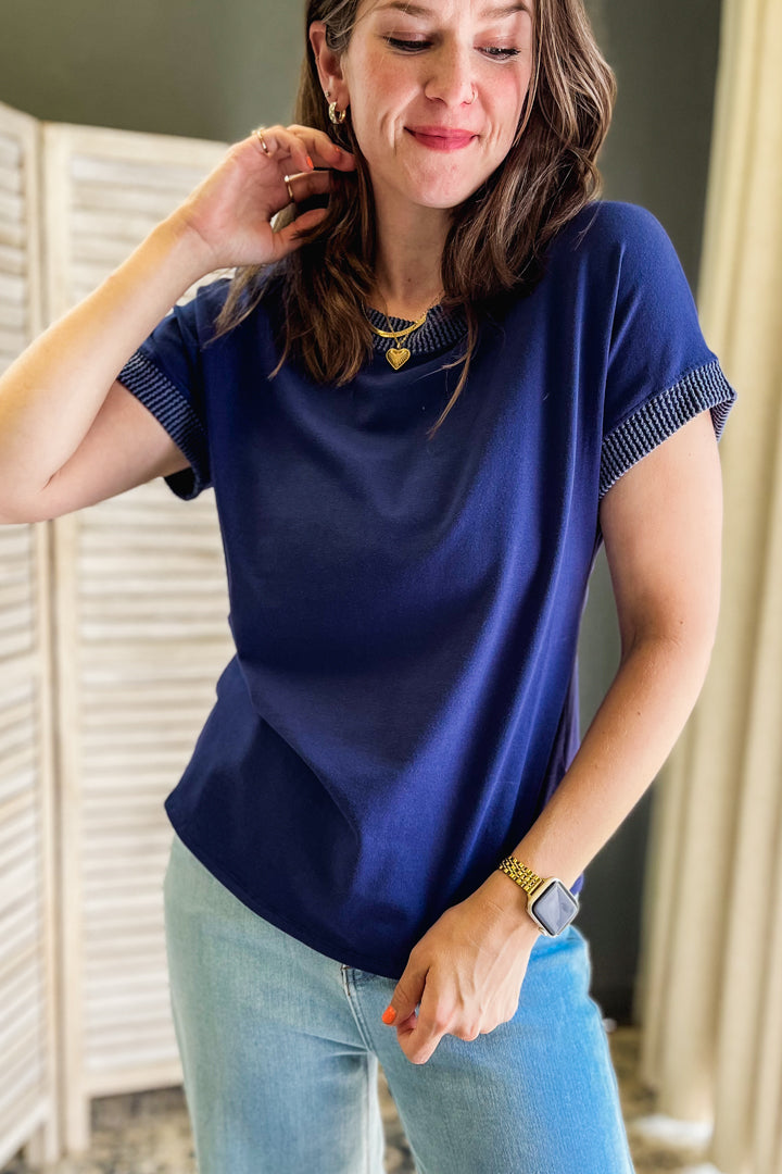 A woman wearing a navy blue short-sleeve top with textured striped trim on the sleeves and neckline, paired with light blue jeans.