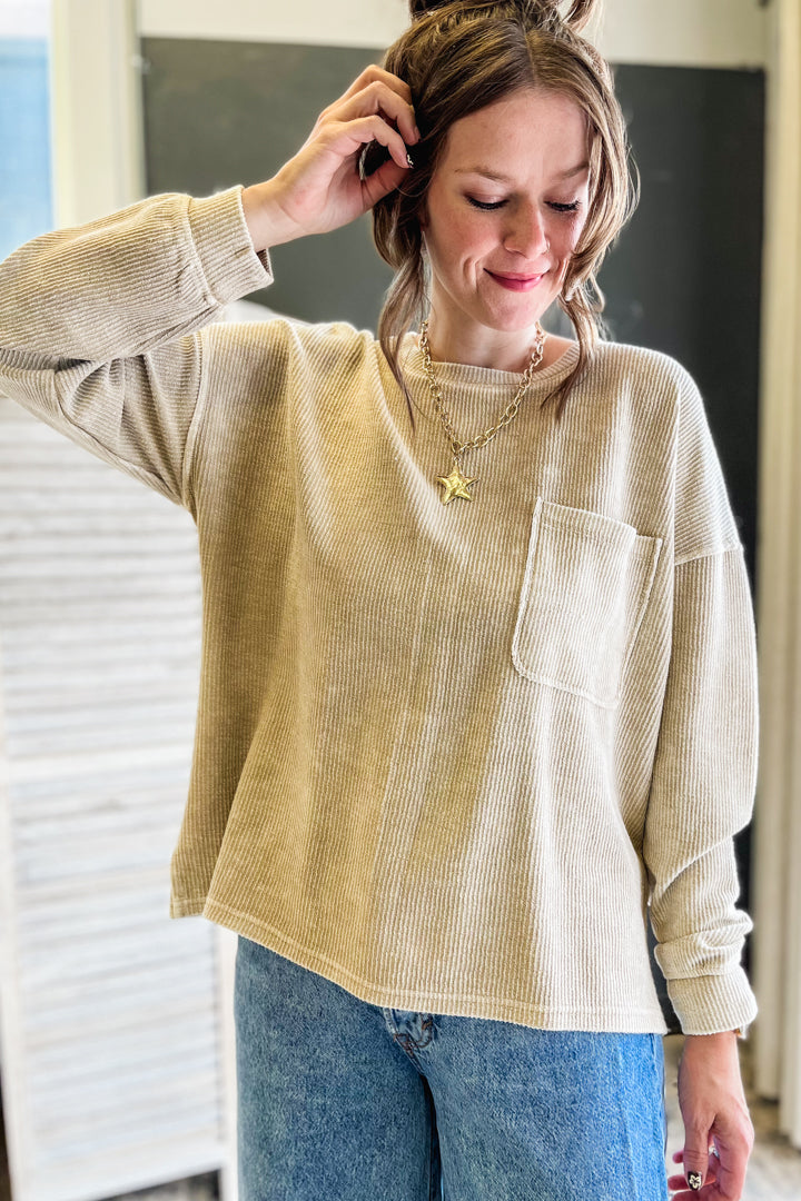 A woman wearing a beige ribbed knit long-sleeve top with a chest pocket, paired with blue jeans and gold necklaces, in an indoor setting.