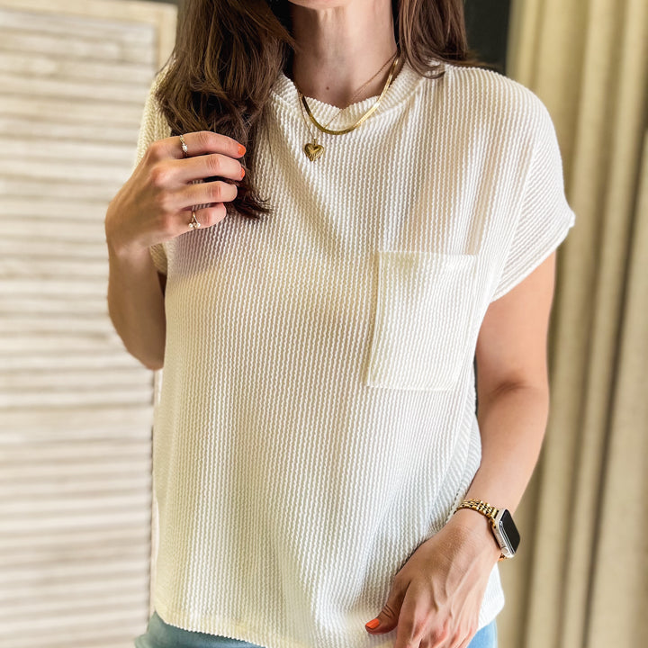 A woman wearing a cream-colored, short-sleeve ribbed knit top with a chest pocket, paired with light blue jeans, standing indoors.