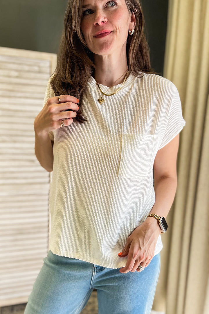 A woman wearing a cream-colored, short-sleeve ribbed knit top with a chest pocket, paired with light blue jeans, standing indoors.
