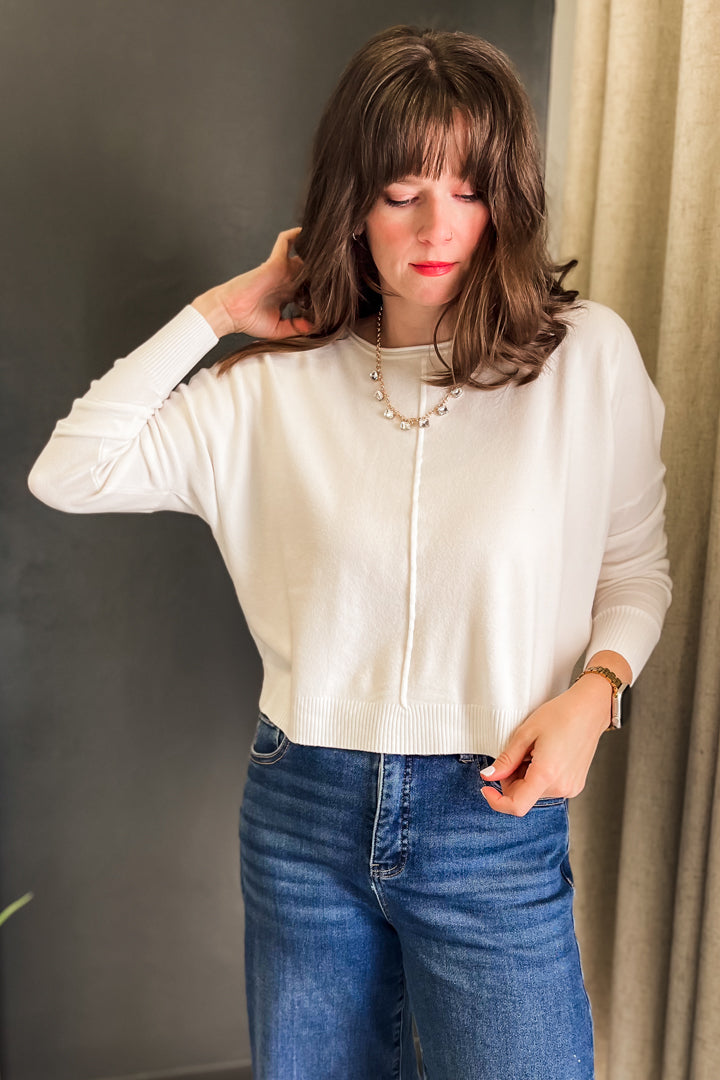 A woman wearing a cream-colored knit zip-up sweater and blue jeans, standing indoors against a dark gray wall and beige curtain.