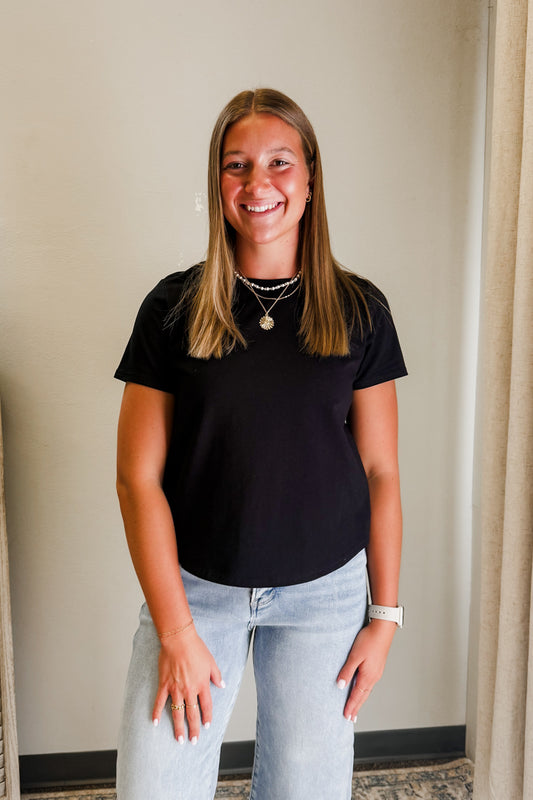 Woman wearing a black t-shirt and light blue jeans standing indoors against a neutral wall.