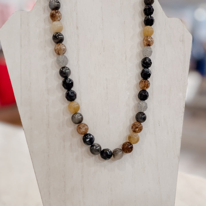 A beaded necklace made of polished round gemstone beads in black, brown, gray, and translucent amber tones, displayed on a white jewelry bust.