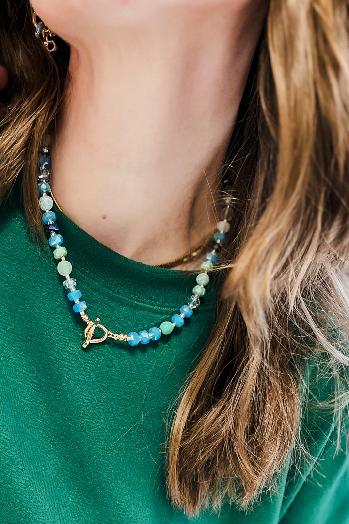 A close-up lifestyle shot of a woman wearing a beaded necklace with light blue, mint green, and white beads, accented by a gold toggle clasp and a thin gold chain.