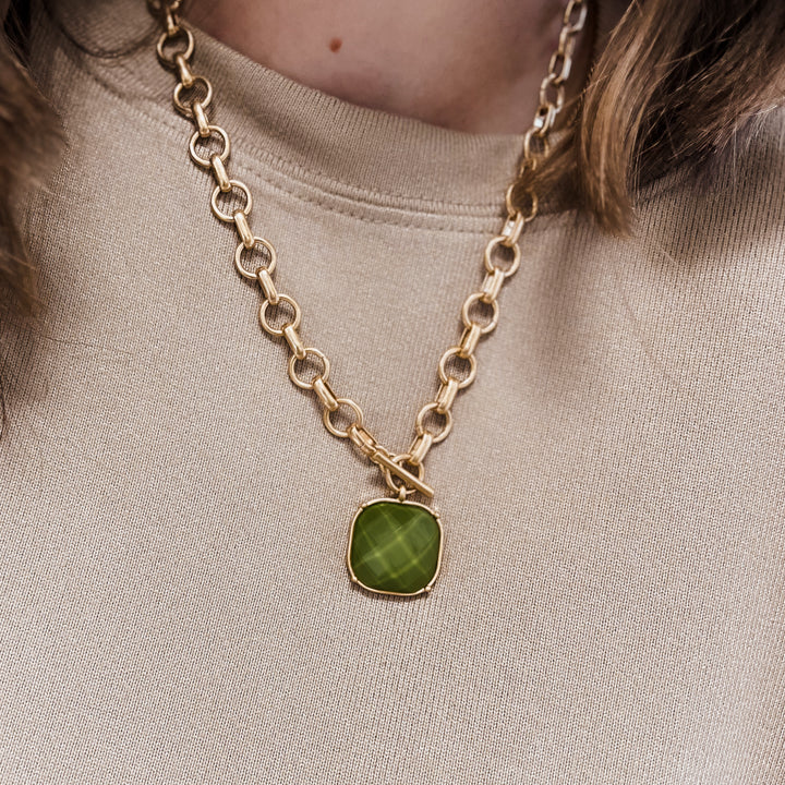 A close-up of a person wearing a gold chain necklace with a cushion-cut green gemstone pendant, paired with a beige crewneck sweatshirt.