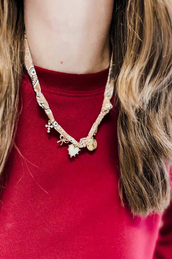 A close-up of a person wearing a gold-toned charm necklace over a red knit top. The necklace features a patterned chain with small dangling charms, including a bow, a flower, and a round charm.