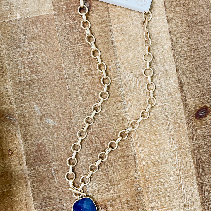 A gold chain necklace with a blue cushion-cut stone pendant, attached to a white paper display card, laid flat on a wooden surface.