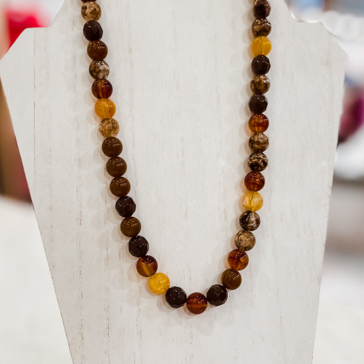 A beaded necklace made of polished round gemstone beads in earthy brown, amber orange, and translucent yellow tones, displayed on a white jewelry bust.