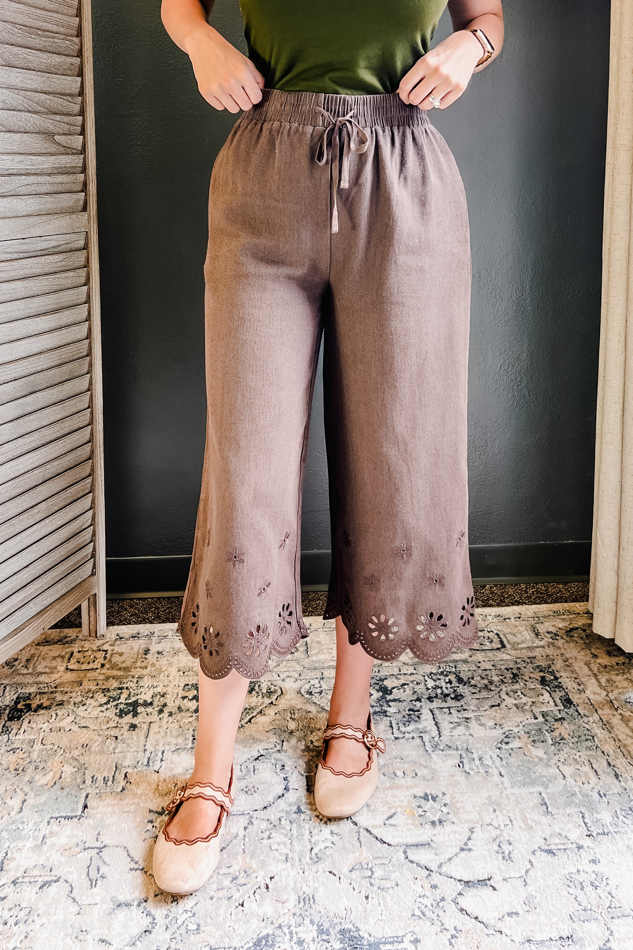 A person wearing loose, wide-leg cropped pants in a muted taupe-brown color with floral eyelet embroidery along the scalloped hems. The pants feature an elastic waistband with a drawstring.