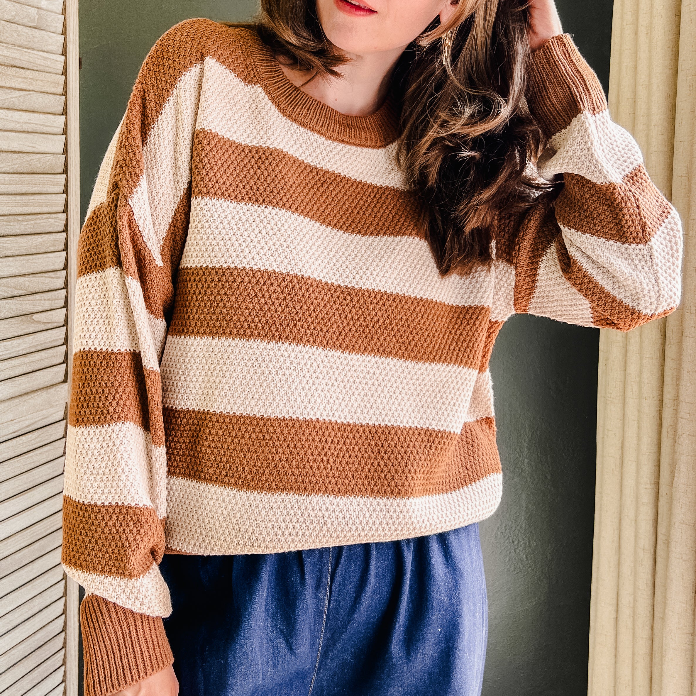 A woman wearing a brown and cream horizontally striped waffle-knit sweater and blue wide-leg pants, standing in an indoor setting with a dark green wall and curtains.