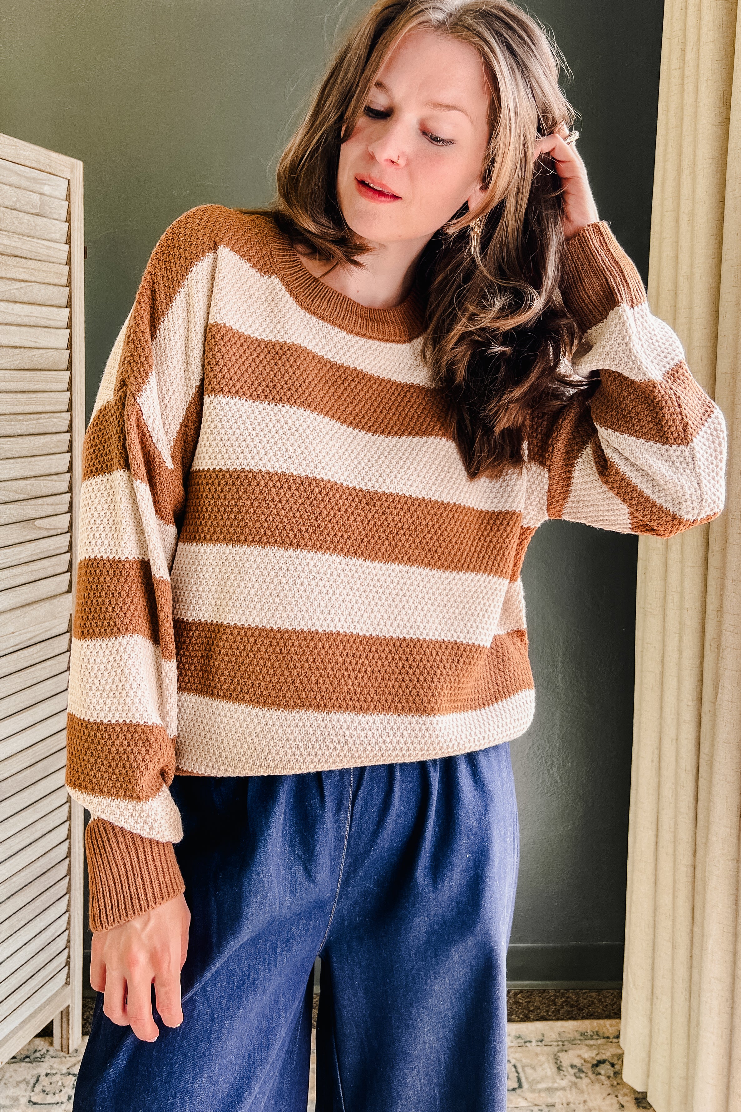 A woman wearing a brown and cream horizontally striped waffle-knit sweater and blue wide-leg pants, standing in an indoor setting with a dark green wall and curtains.
