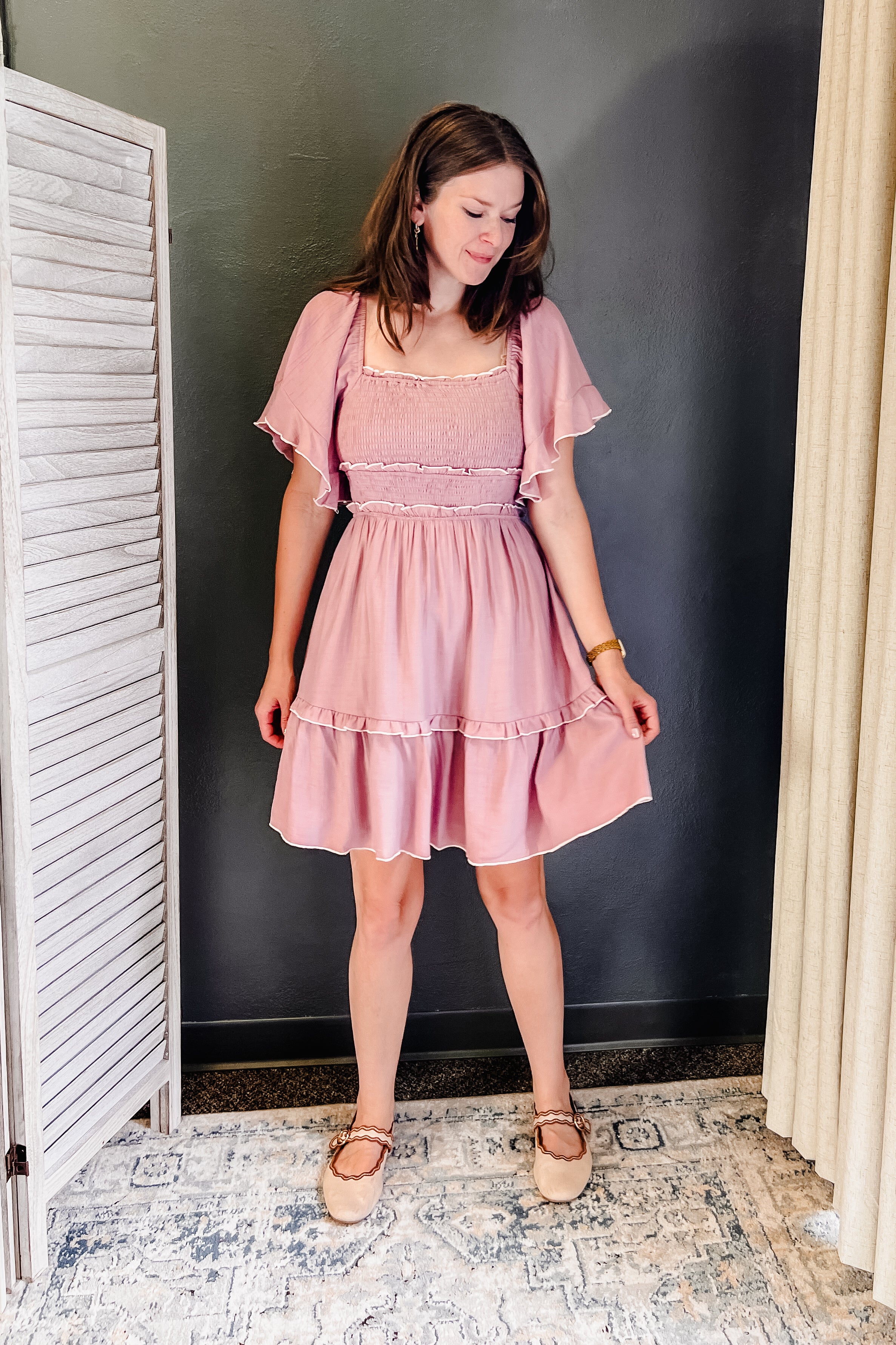 A woman wearing a light pink tiered mini dress with flutter sleeves and ruffle trim, standing in a room with a dark gray wall, white shutter, and patterned rug.