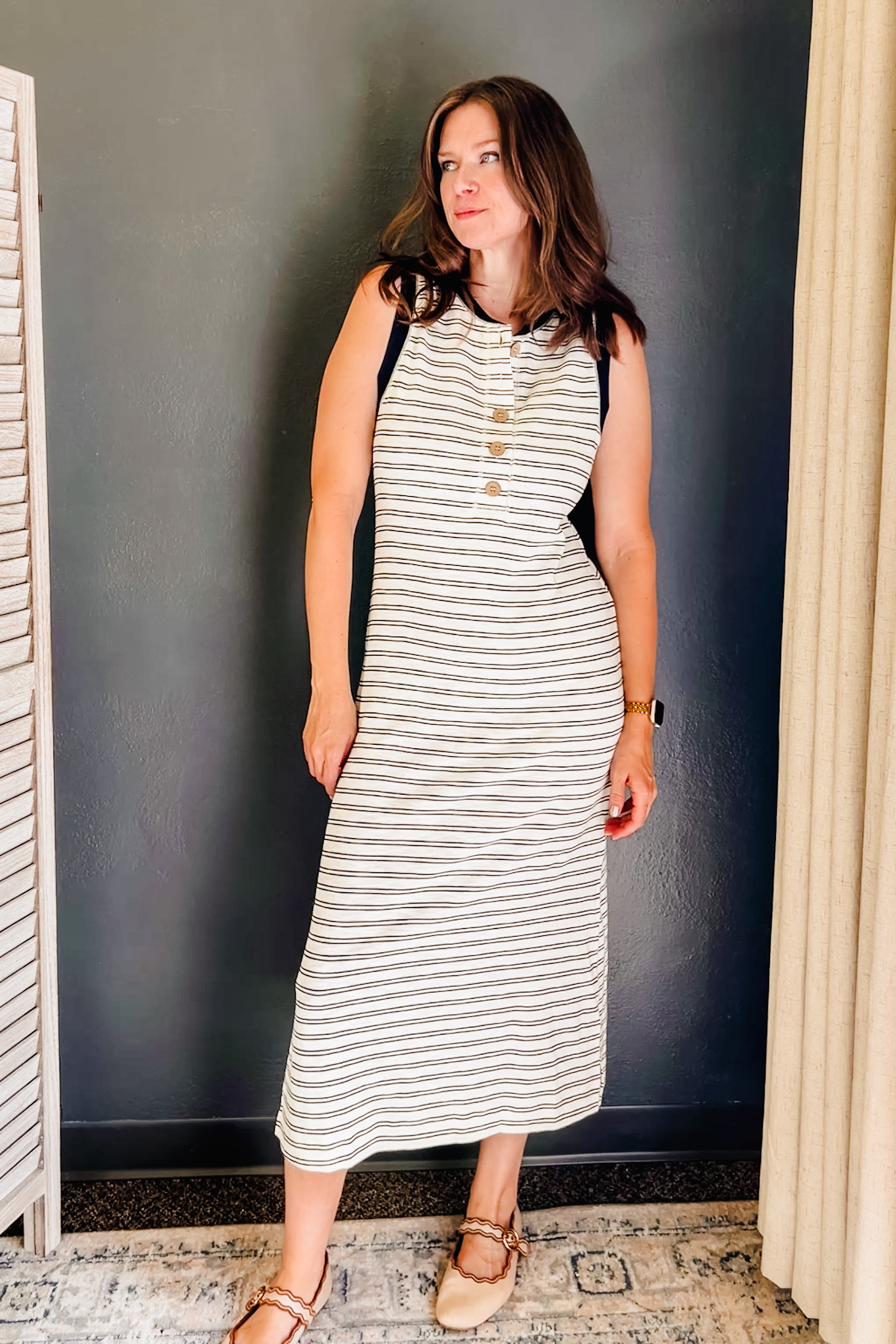A woman modeling a sleeveless midi dress with horizontal navy and off-white stripes, featuring a buttoned henley neckline and black shoulder panels, standing against a dark gray wall.