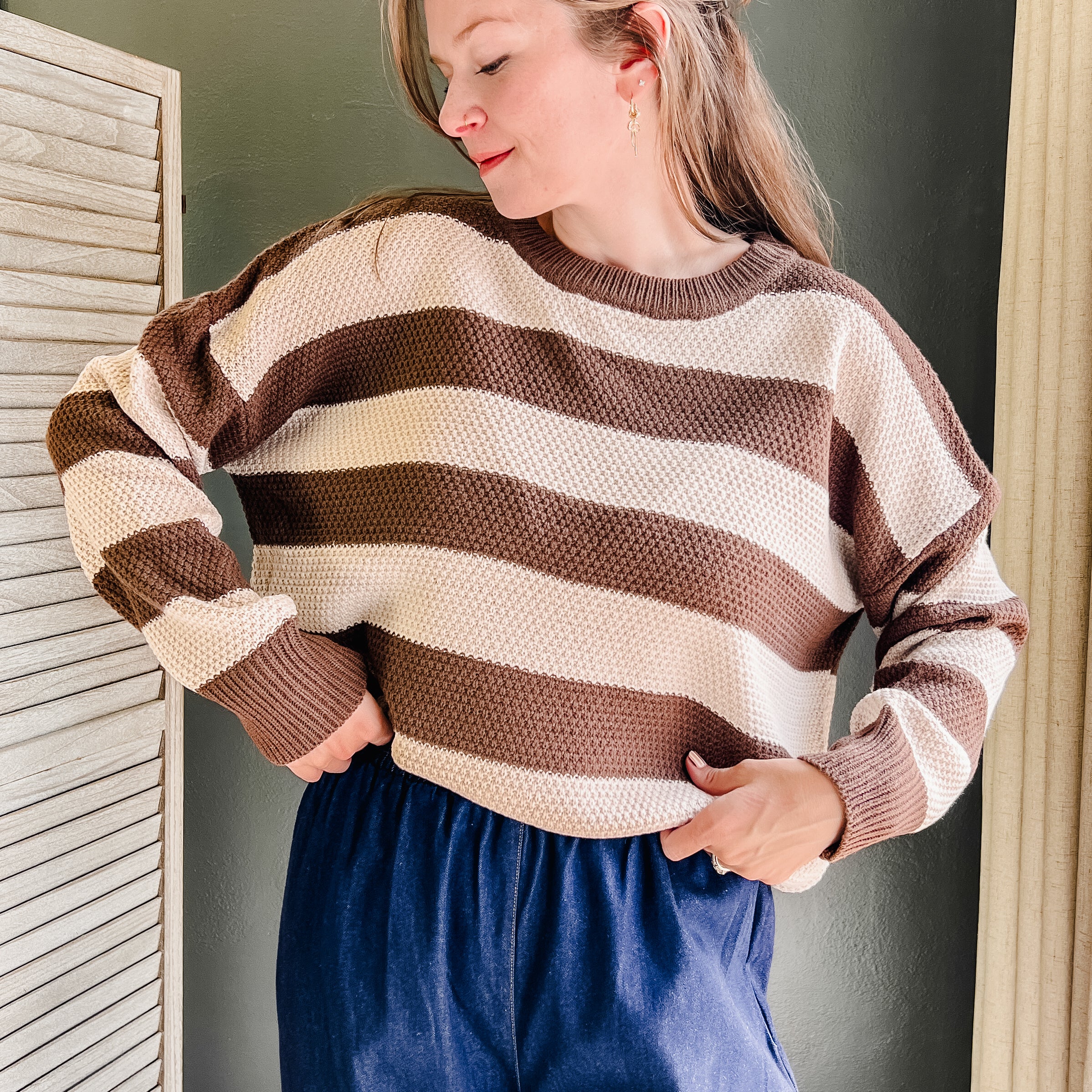 A woman wearing a brown and cream striped knit sweater and blue pants, standing indoors against a dark gray wall.