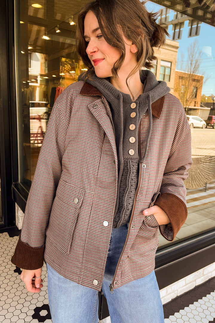 A woman wearing a brown and beige houndstooth jacket with brown ribbed trim over a gray button-front top and light blue jeans, standing in front of a storefront window.