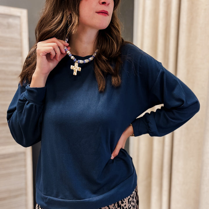A woman wearing a navy blue long-sleeve top and a leopard print skirt, accessorized with a beaded necklace featuring a gold cross pendant, posing in front of a neutral indoor background.