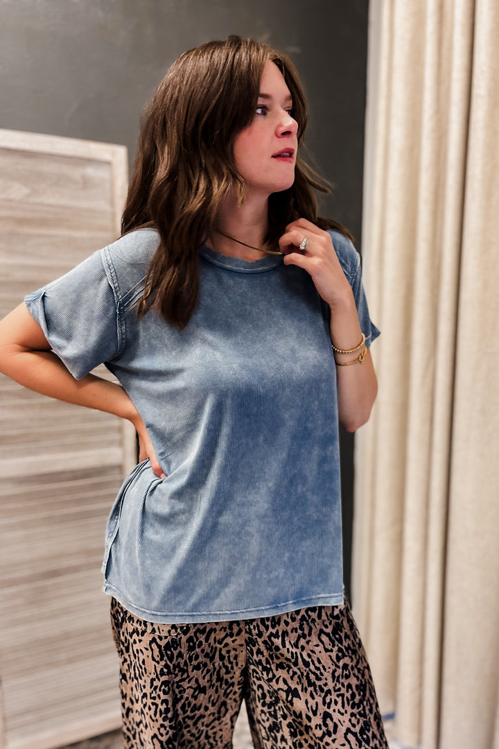 A woman wearing a light blue acid-wash short-sleeve t-shirt and leopard print pants, standing in an indoor setting with a gray wall and beige curtains.