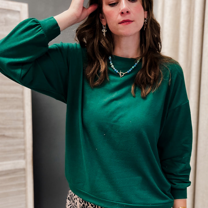 A woman wearing a dark green long-sleeve crewneck sweatshirt and a leopard print skirt, standing in front of a neutral indoor background.