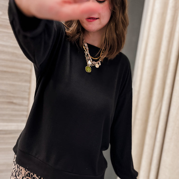 A woman wearing a black long-sleeve crewneck sweatshirt and a beige leopard print skirt, with layered gold necklaces, posing indoors with her hand partially covering the camera.