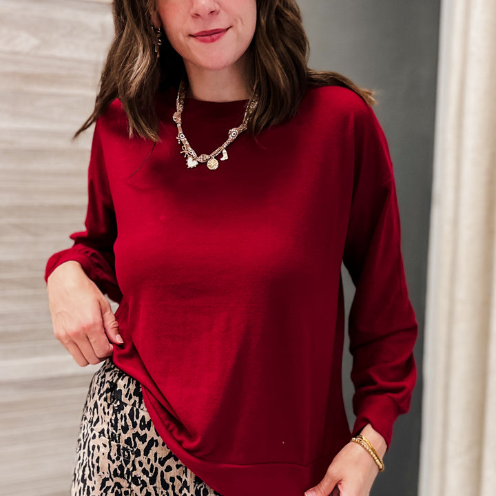 A woman wearing a long-sleeve crewneck sweatshirt in deep red, paired with a beige and black leopard print skirt, standing indoors.