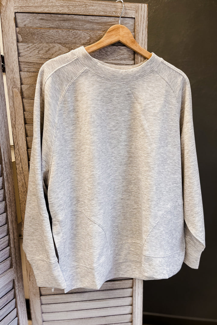 A light heather gray crewneck sweatshirt hanging on a wooden hanger against a rustic wooden slat door.