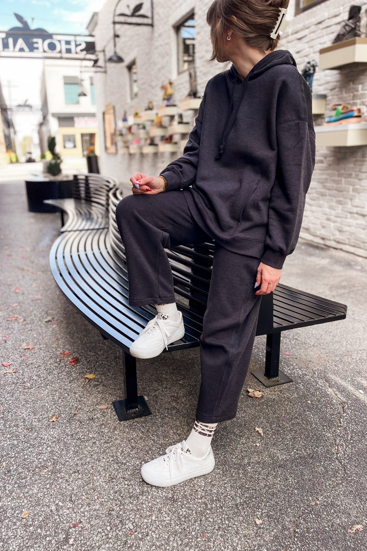 A person wearing a dark charcoal gray matching lounge set consisting of a pullover hoodie and wide-leg sweatpants, sitting on a curved black bench in an outdoor urban setting.