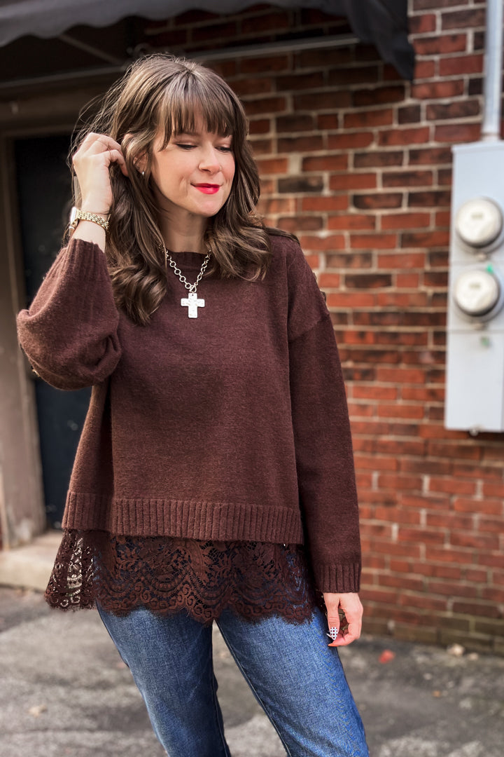 A woman wearing a brown knit sweater with a lace hem, blue jeans, and a silver cross necklace, standing outdoors in front of a brick wall.