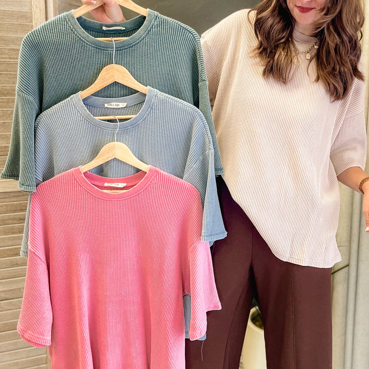 A woman stands indoors holding three ribbed knit short-sleeve tops on wooden hangers, shown in a lifestyle setting. The tops are in three different colors: dusty blue, light blue, and pink, layered on a neutral background.