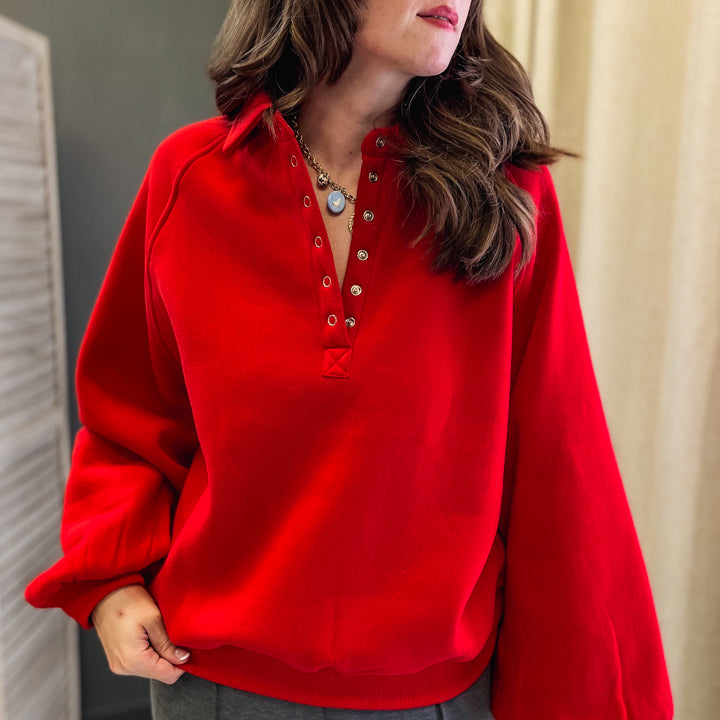 A woman wearing a bright red long-sleeve henley-style top with a buttoned collar and gray pants, standing indoors against a neutral curtain and wall background.