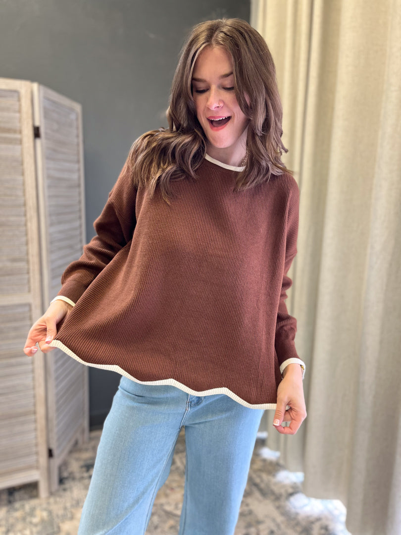 A woman wearing a brown knit sweater with cream ribbed trim at the crew neck, cuffs, and scalloped hem, paired with light blue jeans in an indoor setting.