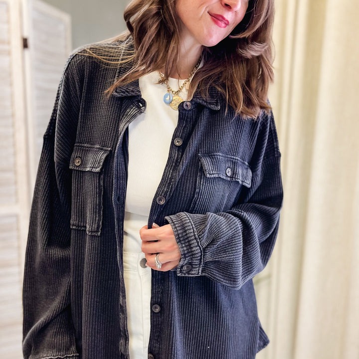 A woman wearing an oversized black corduroy button-up shirt jacket over a white top and white pants, standing indoors against a neutral beige curtain background.
