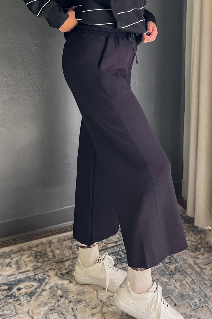 A person wearing black wide-leg cropped pants with a drawstring waist, paired with white sneakers and socks, standing in an indoor setting with a patterned rug and gray wall.